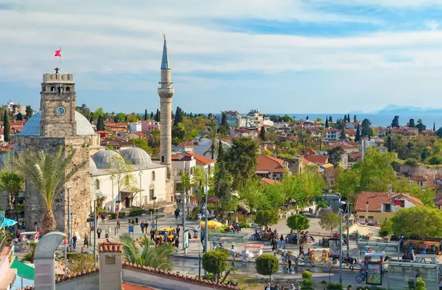 Antalya's 19th-century clock tower crowned by a Turkish flag stands beside the domed Tekeli Mehmet Pasha Mosque and its slender minaret, overlooking the busy main square of Kaleici old town with the blue Mediterranean glinting on the horizon.