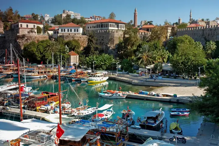 Kaleici harbor in Antalya tightly packed with colorful traditional wooden fishing boats and excursion vessels in vivid turquoise water, with the slender Yivli Minaret and palm-lined quays rising above the stone waterfront wall.