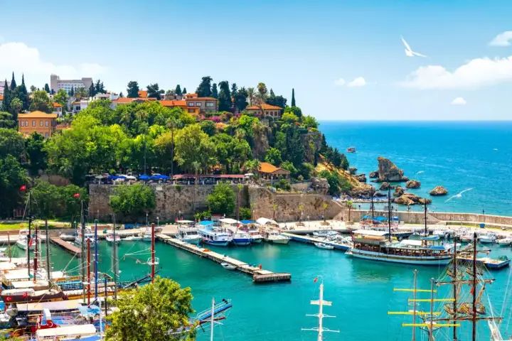 La marina del Kaleici de Antalya en un brillante día de verano, con goletas tradicionales de mástiles altos amarradas a muelles de piedra, la ladera verde arbolada del casco antiguo elevándose sobre murallas medievales y una gaviota solitaria planeando sobre el Mediterráneo abierto.