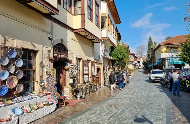 Una via commerciale acciottolata del Kaleici di Antalya dove la parete esterna di un edificio ottomano in pietra è coperta dall'alto in basso di piatti in ceramica turca dipinti a mano con motivi geometrici in blu, rosso e verde, con altra ceramica esposta su un basso tavolo all'esterno.