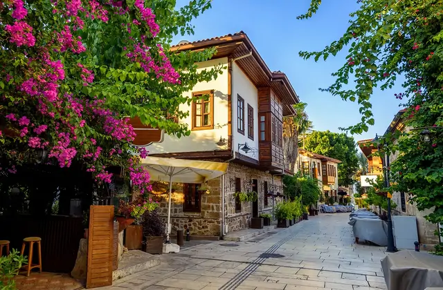Una esquina del Kaleici de Antalya donde una explosión de buganvilla de color magenta intenso cuelga sobre una mansión otomana de dos plantas bellamente restaurada, construida en piedra tallada a nivel de calle con pisos superiores de madera, con un café de terraza bajo una sombrilla crema.