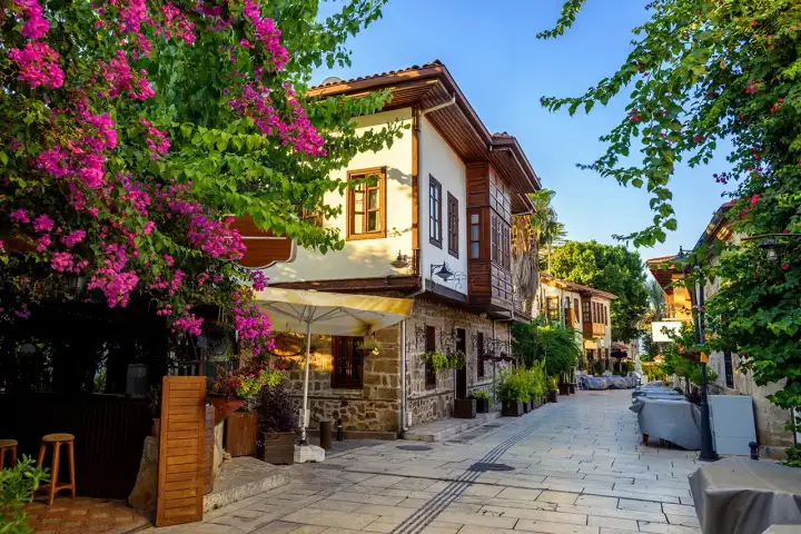 Una esquina del Kaleici de Antalya donde una explosión de buganvilla de color magenta intenso cuelga sobre una mansión otomana de dos plantas bellamente restaurada, construida en piedra tallada a nivel de calle con pisos superiores de madera, con un café de terraza bajo una sombrilla crema.