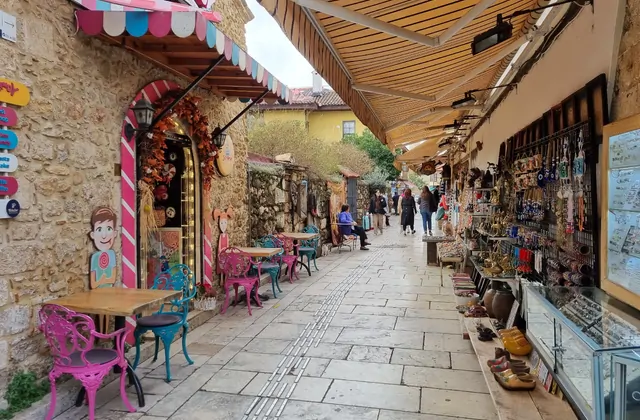 Un stretto corridoio del mercato coperto all'interno del bazar del Kaleici di Antalya, con audaci tende a righe in cima, vivaci sedie da caffè in ghisa rosa raggruppate a sinistra accanto a un ingresso di negozio a righe di caramella e bancarelle di souvenir con gioielli e amuleti nazar a destra.