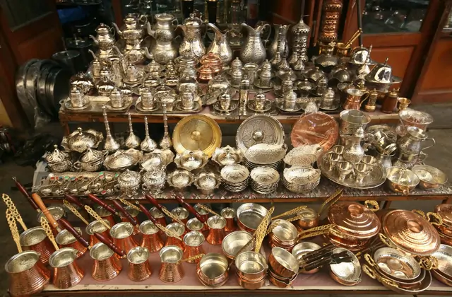 A market stall table in Gaziantep's copper bazaar overflows with densely packed hand-engraved silverware — ornate teapots, ladles, sugar bowls and small ibrik coffee pots arranged in rows, their polished surfaces gleaming under ambient light.