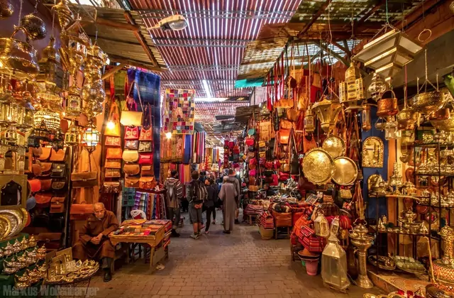 Un stretto corridoio del bazar di Gaziantep si allunga in profondità con lanterne e piatti di rame appesi in alto, tessuti colorati che scendono da entrambi i lati e acquirenti che si muovono tra bancarelle piene di artigianato, borse e lavori in metallo.