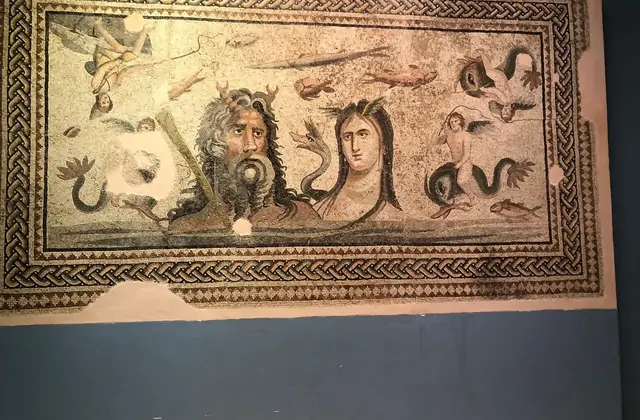 A large Roman mosaic panel at Gaziantep's Zeugma Mosaic Museum depicts the sea deities Oceanus and Tethys — a bearded male face with horns flanks a serene female portrait, surrounded by dolphins, fish and a Cupid figure within a braided geometric border.