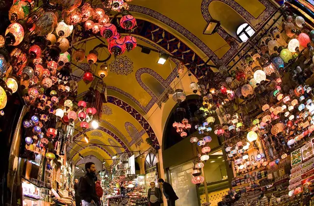 Centinaia di lanterne turche a mosaico dipinte a mano in rosso, blu, verde e ambra affollano il soffitto ad arco in pietra di una sala del bazar di Gaziantep, proiettando luci dai toni preziosi lungo un passaggio fiancheggiato da negozi di souvenir sotto arcate ottomane.