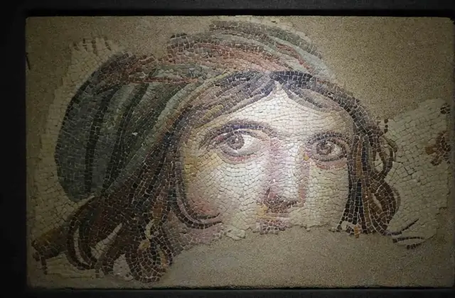 L'iconico mosaico della Ragazza Gitana da Zeugma romano è esposto su sfondo scuro al Museo del Mosaico di Zeugma di Gaziantep — il suo sguardo diretto e malinconico reso in sottili tessere ocra, grigie e rosa, con i capelli scuri ondulati che incorniciano un volto senza tempo.