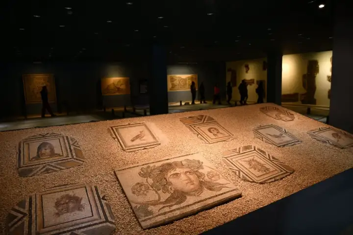 The dimly lit main gallery of Gaziantep's Zeugma Mosaic Museum features a large illuminated floor display of Roman portrait mosaics — multiple medallion panels with faces and birds set in sandy gravel, with silhouetted visitors moving among other mosaics in the background.