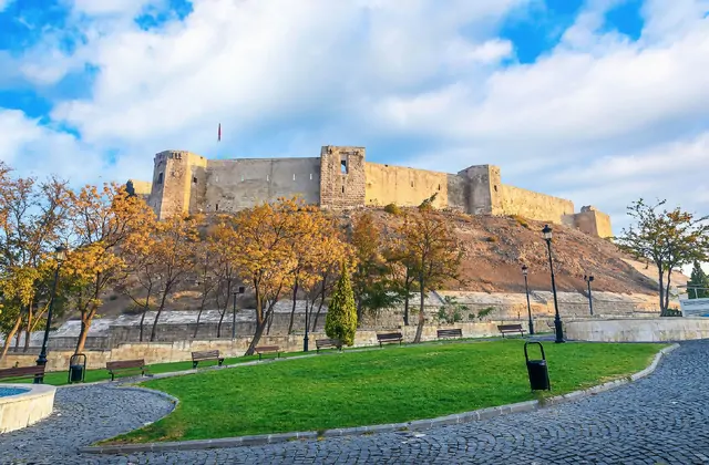 Il Castello di Gaziantep siede sulla sua altura rocciosa in autunno, le sue torri in pietra e le mura merlate incorniciate da alberi dalle foglie dorate e una passeggiata in acciottolato sottostante, con una bandiera turca visibile in cima e un cielo velato sopra.