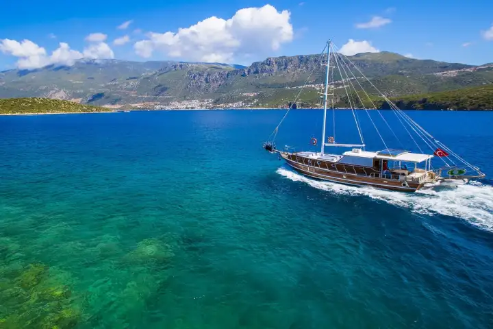 Seen from a low aerial angle, a wooden gulet flying the Turkish flag cuts across water that grades from vivid turquoise shallows to deep blue, with a white coastal town spread across rugged mountain slopes on the Turkish Aegean shore.