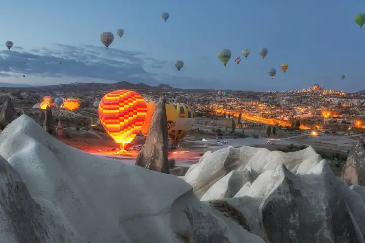At blue hour, ground teams inflate hot air balloons against a deep twilight sky while the lights of a Cappadocian town glow below, Turkey, marking the start of the early morning ritual.