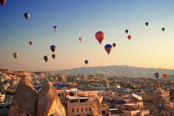 The morning sky above a Cappadocian cave village, Turkey, fills with dozens of ascending hot air balloons, their reflections catching the first warm light as the valley wakes.