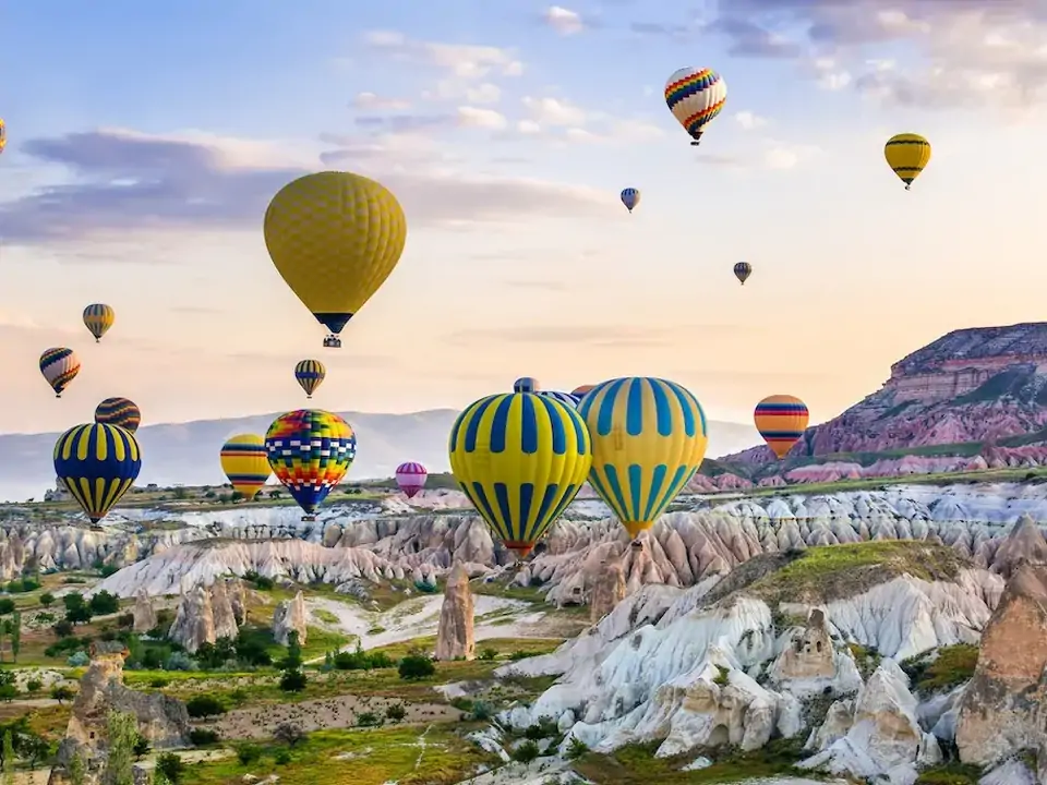 Mongolfiere colorate sorvolano la Valle delle Rose in Cappadocia, Turchia, dove le calde sfumature rosa e ambra delle formazioni rocciose erose si intensificano nella morbida luce del mattino.
