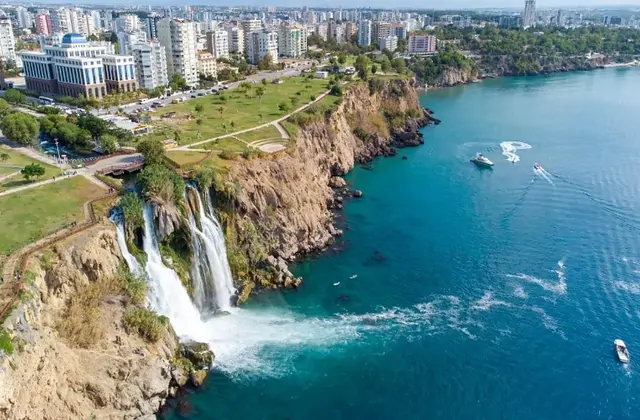 Aerial drone view of Lower Düden Waterfall in Antalya showing the cascade tumbling from the rocky clifftop park platform down to the sea, with multiple pleasure boats cutting white wakes through turquoise water and the dense city grid stretching inland.