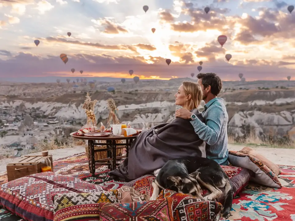 A couple and their dog enjoy an outdoor picnic breakfast in Cappadocia, Turkey, as hot air balloons drift peacefully overhead through the early morning sky.