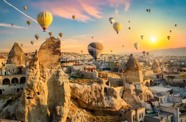La calda luce dorata del mattino cade sulla facciata in pietra di un hotel grotta della Cappadocia, Turchia, mentre alcune mongolfiere derivano nel cielo luminoso in lontananza.
