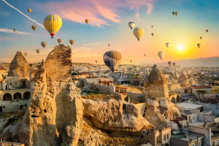 Warm golden morning light falls on the stone facade of a Cappadocian cave hotel, Turkey, while several hot air balloons drift across the glowing sky in the distance.