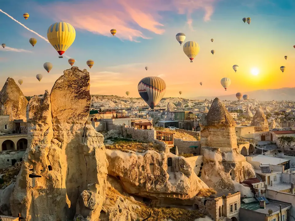 La calda luce dorata del mattino illumina la facciata in pietra di un hotel grotta in Cappadocia, Turchia, mentre diversi palloni aerostatici scivolano nel cielo luminoso in lontananza.