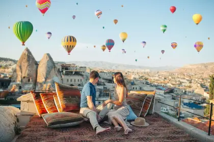 A couple relaxes on a carved stone terrace in Cappadocia, Turkey, as a sky crowded with colorful hot air balloons passes overhead in the gentle morning breeze.
