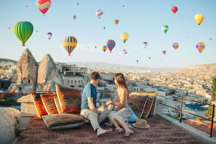 Una pareja descansa en una terraza de piedra tallada en Capadocia, Turquía, mientras un cielo repleto de coloridos globos aerostáticos pasa sobre ellos en la suave brisa matinal.