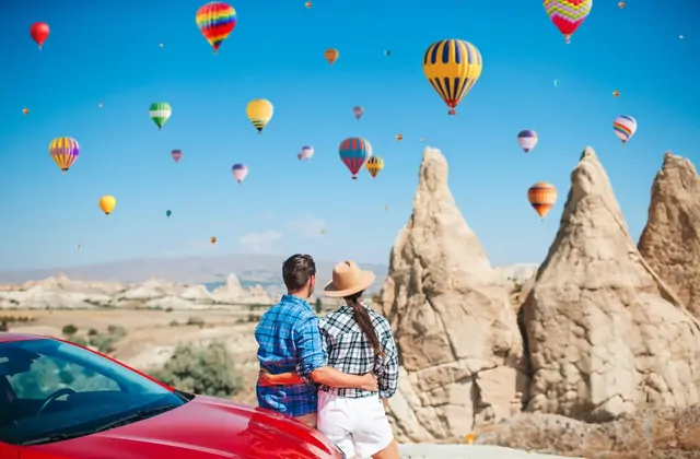 Una coppia posa per una foto di viaggio accanto alle guglie di roccia vulcanica in Cappadocia, Turchia, con alcune vivaci mongolfiere visibili nel cielo azzurro sopra il terreno accidentato.