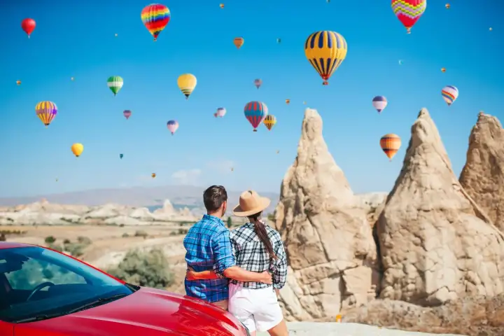 A couple poses for a roadtrip photo beside volcanic rock spires in Cappadocia, Turkey, with several bright hot air balloons visible in the blue sky above the rugged terrain.