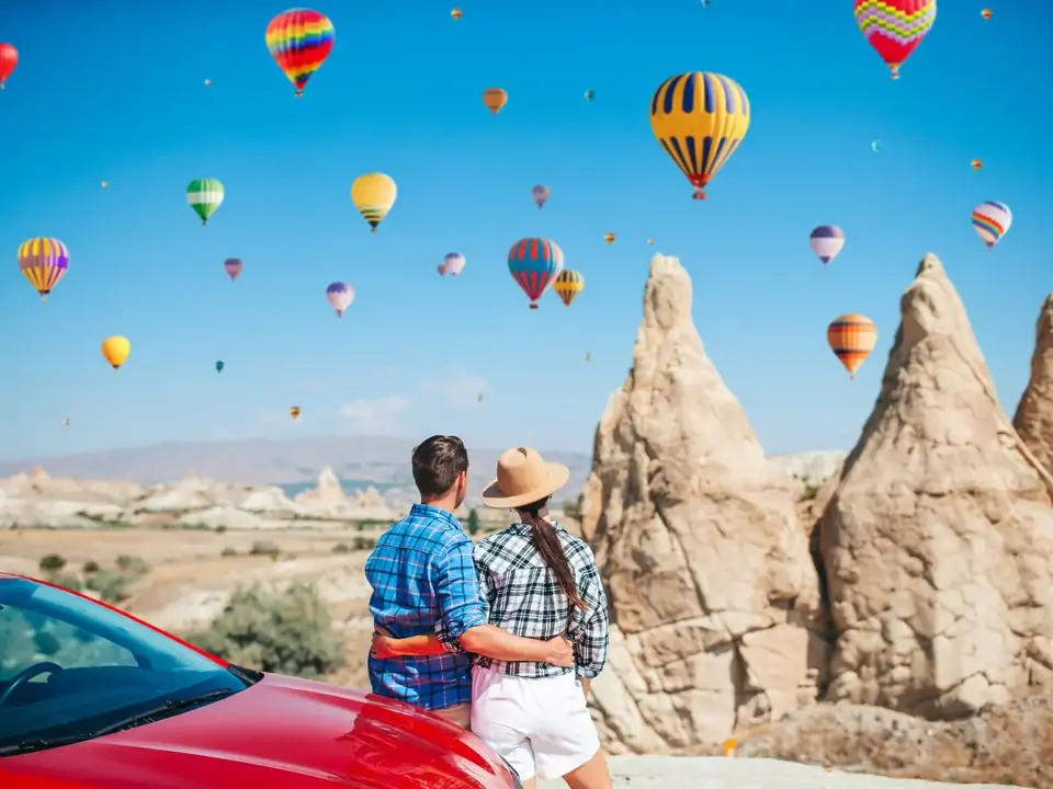 Una pareja posa para una foto de viaje por carretera junto a agujas de roca volcánica en Capadocia, Turquía, con varios globos aerostáticos brillantes visibles en el cielo azul sobre el paisaje rocoso.