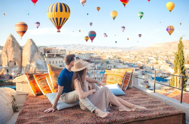Two people unwind on a terrace carved into the soft volcanic rock of a Cappadocian cave hotel, Turkey, with hot air balloons painting the open sky beyond the stone railing.