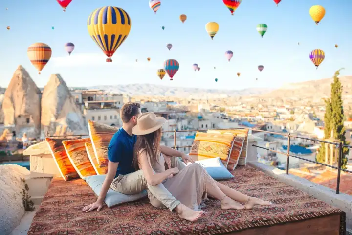 Two people unwind on a terrace carved into the soft volcanic rock of a Cappadocian cave hotel, Turkey, with hot air balloons painting the open sky beyond the stone railing.