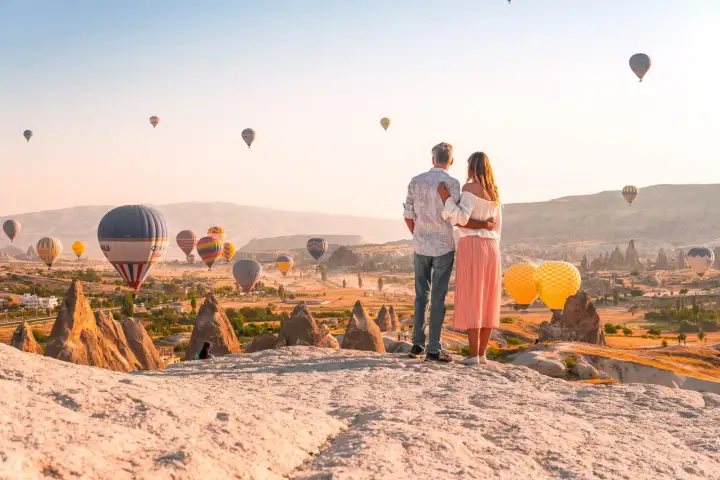 Una coppia si trova in un belvedere elevato con vista sulle valli della Cappadocia, Turchia, all'alba mentre il cielo dietro di loro si riempie di mongolfiere in ascesa che brillano nella prima luce.
