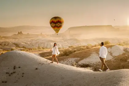 A couple walks hand in hand through Golden Valley in Cappadocia, Turkey, with a single large hot air balloon floating low overhead against the canyon's warm amber walls.