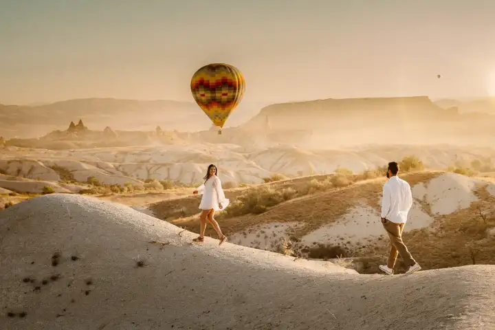 Una pareja camina de la mano por el Valle Dorado en Capadocia, Turquía, con un único gran globo aerostático flotando bajo sobre sus cabezas junto a las cálidas paredes ámbar del cañón.