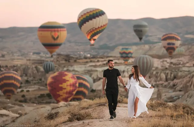 Due figure camminano attraverso una valle della Cappadocia, Turchia, rimpicciolite da enormi mongolfiere che passano molto vicine sopra di loro, i loro massicci involucri che oscurano una sezione del cielo.