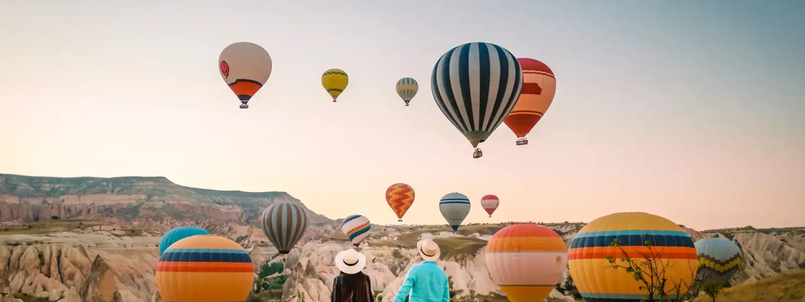 Una coppia passeggia insieme attraverso una pittoresca valle della Cappadocia, Turchia, mentre una sfilata di mongolfiere dai colori vivaci si muove silenziosamente nel cielo sopra il paesaggio rupestre.