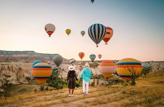 A couple strolls together through a scenic Cappadocian valley, Turkey, while a parade of hot air balloons in vivid colors moves silently through the sky above the rock-cut landscape.