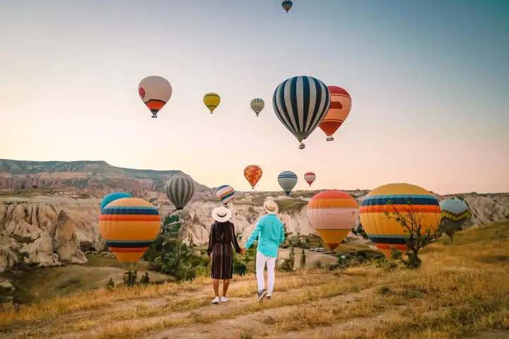 A couple strolls together through a scenic Cappadocian valley, Turkey, while a parade of hot air balloons in vivid colors moves silently through the sky above the rock-cut landscape.