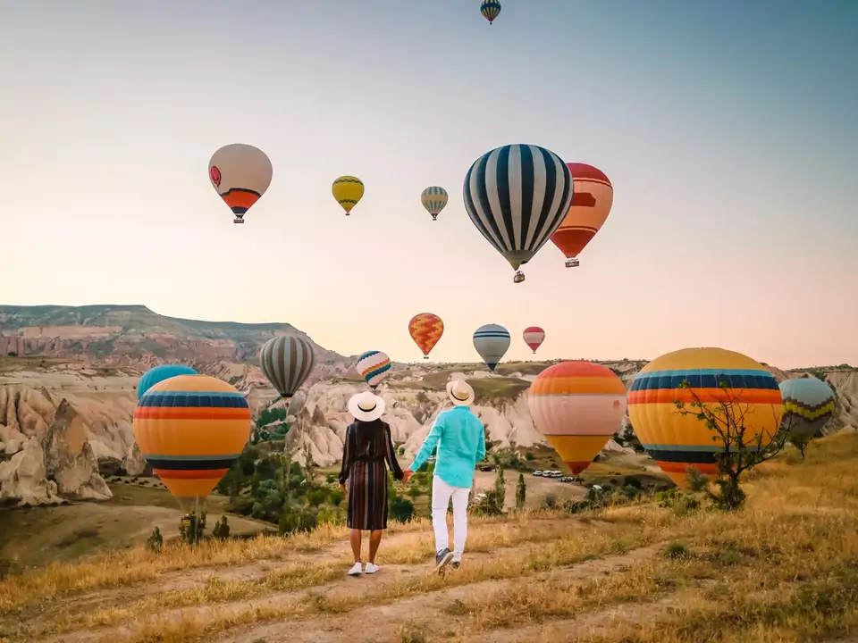 Una coppia passeggia insieme in una valle scenografica della Cappadocia, in Turchia, mentre una serie di mongolfiere colorate si muove in silenzio sopra il paesaggio scavato nella roccia.