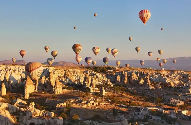 Alti camini di fate si innalzano dall'altopiano della Cappadocia, Turchia, mentre il cielo mattutino sopra è vivo di decine di mongolfiere a varie altitudini che derivano nell'aria ferma.