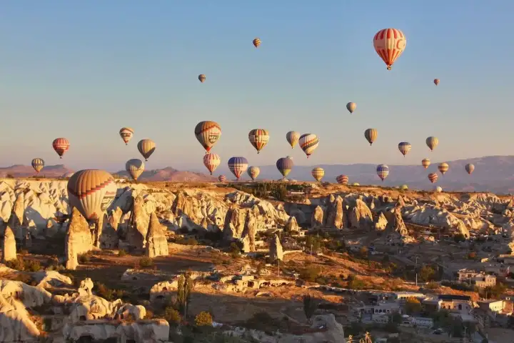 Tall fairy chimneys rise from the Cappadocian plateau, Turkey, while the morning sky above is alive with dozens of hot air balloons at various altitudes drifting in the still air.