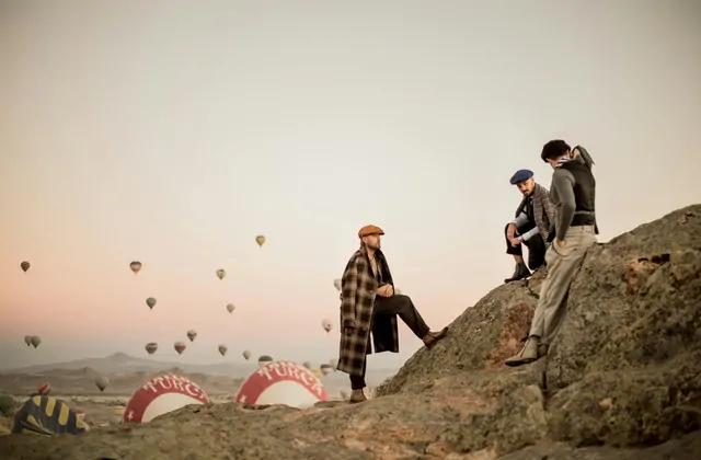 A fashion portrait taken on a Cappadocian rock ridge, Turkey, with hot air balloons floating gracefully in the distance behind the subject against a clear blue sky.