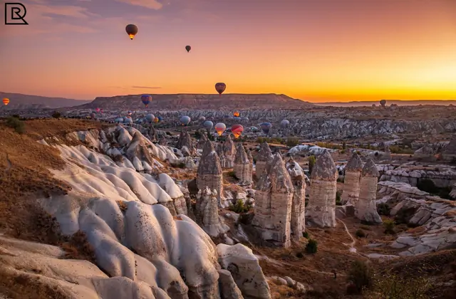 I pilastri di roccia fallici della Valle dell'Amore in Cappadocia, Turchia, si stagliano contro un cielo al tramonto arancione intenso mentre alcune mongolfiere si librano sulla valle al crepuscolo.