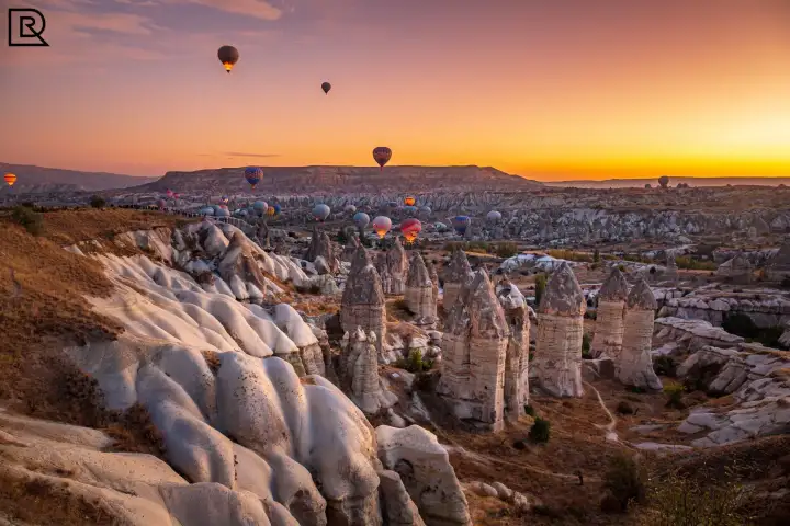 I pilastri di roccia fallici della Valle dell'Amore in Cappadocia, Turchia, si stagliano contro un cielo al tramonto arancione intenso mentre alcune mongolfiere si librano sulla valle al crepuscolo.