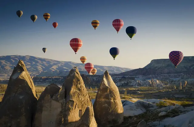 Un amplio panorama matinal sobre un valle de chimeneas de hadas en Capadocia, Turquía, captura decenas de globos aerostáticos a múltiples alturas dispersos por el vasto cielo abierto.