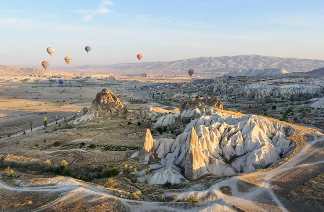 Una valle aperta della Cappadocia si estende ampia sotto un cielo mattutino, Turchia, con mongolfiere sparse sull'orizzonte come gioielli dispersi sul terreno di roccia ocra e grigia.