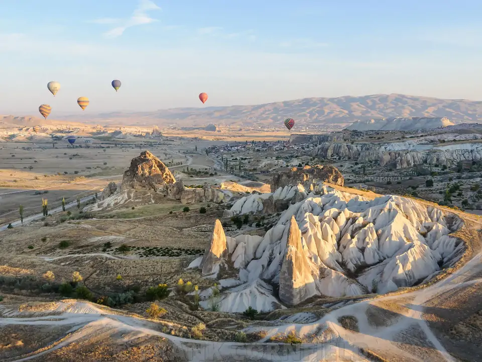 Un’ampia valle della Cappadocia si estende sotto il cielo del mattino, con mongolfiere sparse all’orizzonte come gioielli sopra il terreno roccioso color ocra e grigio.