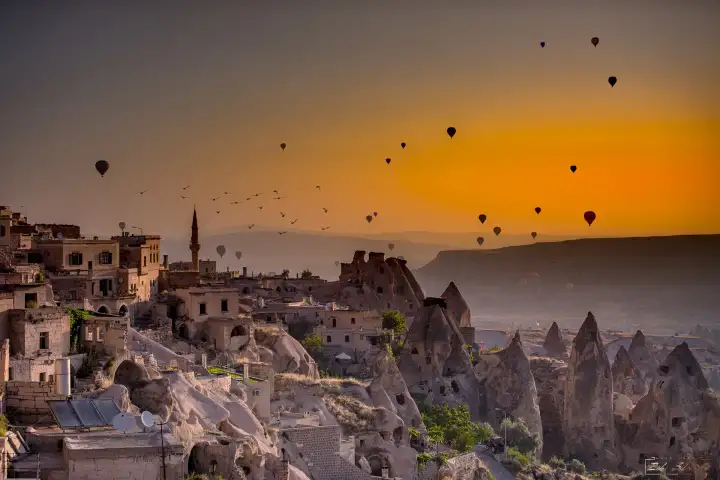 An intense orange dawn sky blazes over a Cappadocian cave village, Turkey, silhouetting the rooftops and chimneys while balloons rise into the vivid sunrise in the distance.