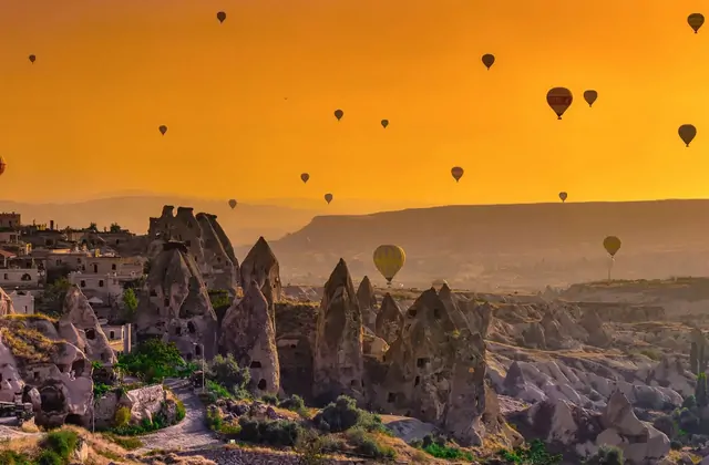 Un cielo naranja ricamente saturado llena el encuadre sobre un pueblo cueva de Capadocia, Turquía, mientras un vuelo en globo aerostático pasa a través del cálido resplandor de la luz de primera hora.