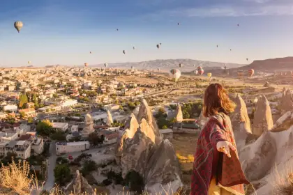 A woman stands at a Göreme viewpoint in Cappadocia, Turkey, arms outstretched as the famous balloon-dotted panorama of the valley unfolds behind her at sunrise.