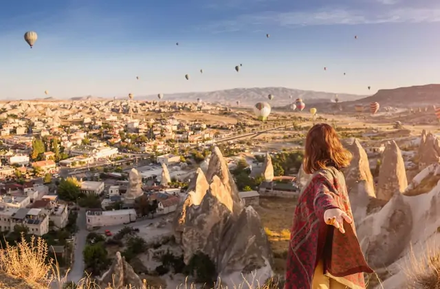 A woman stands at a Göreme viewpoint in Cappadocia, Turkey, arms outstretched as the famous balloon-dotted panorama of the valley unfolds behind her at sunrise.